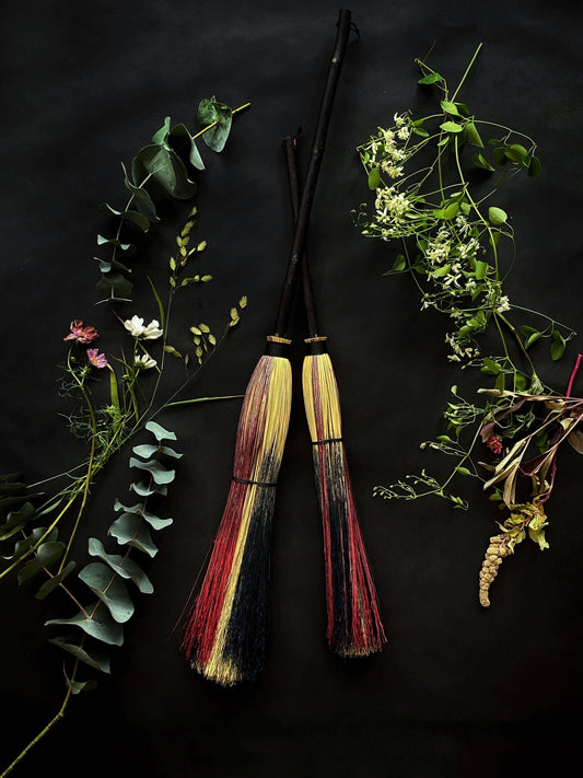 Two broomsticks with multicolored handles surrounded by greenery on a dark background