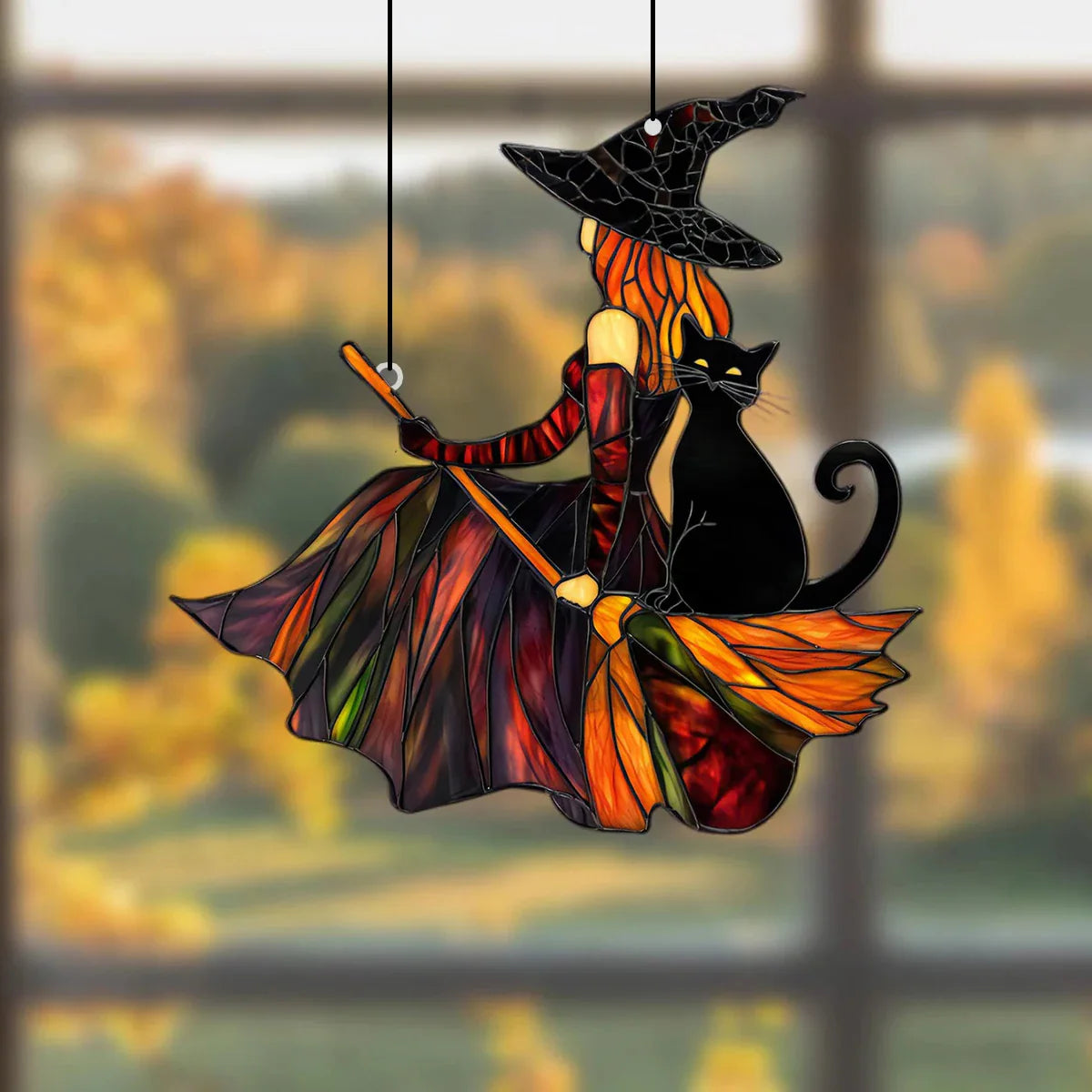 Decorative acrylic stained glass witch and black cat on a broom hanging against a blurred autumn window background.