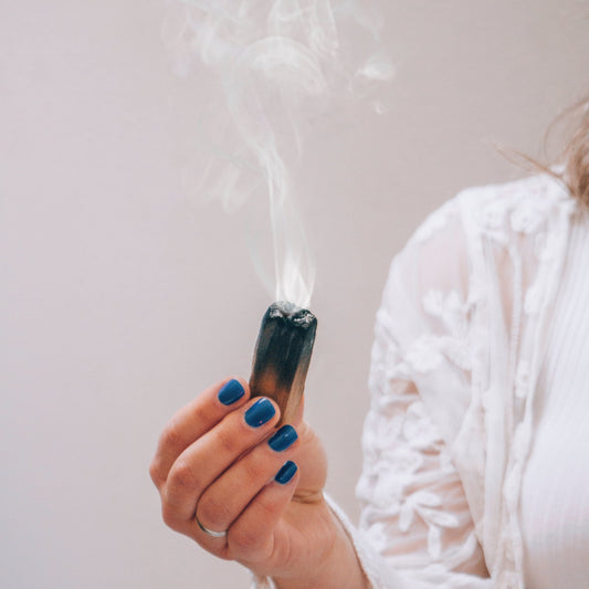 A hand with blue painted nails holding a smoking resin bundle against a white background