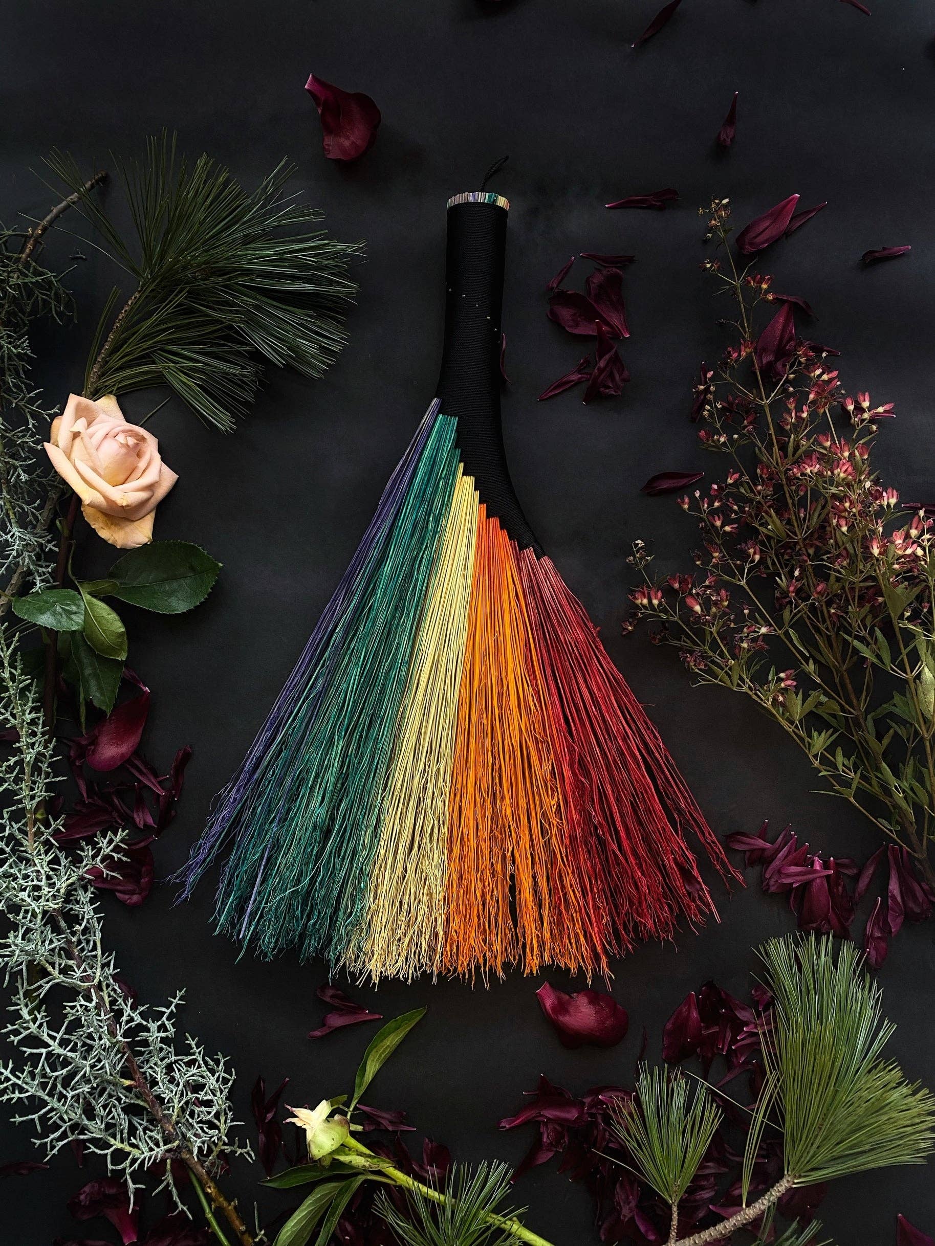 Colorful broom with rainbow bristles on a dark background with flowers and plants.